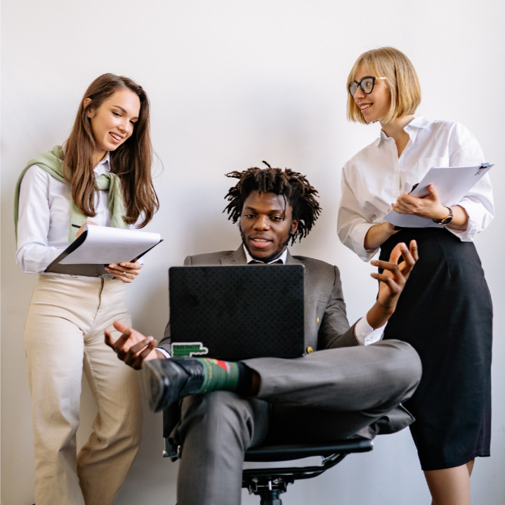Three people in a professional setting with one person using a laptop, another holding a tablet, and a third person standing.
