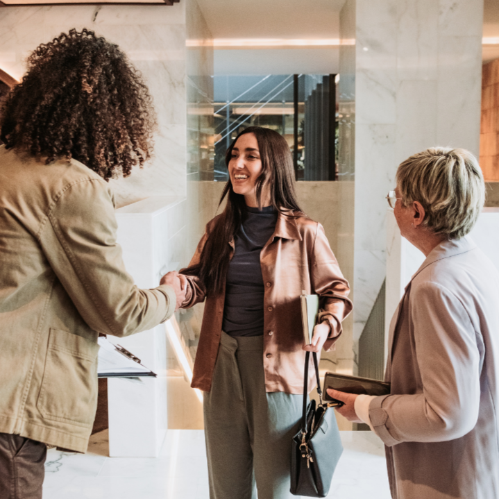 Two women shaking hands in a modern indoor setting with a third woman observing.