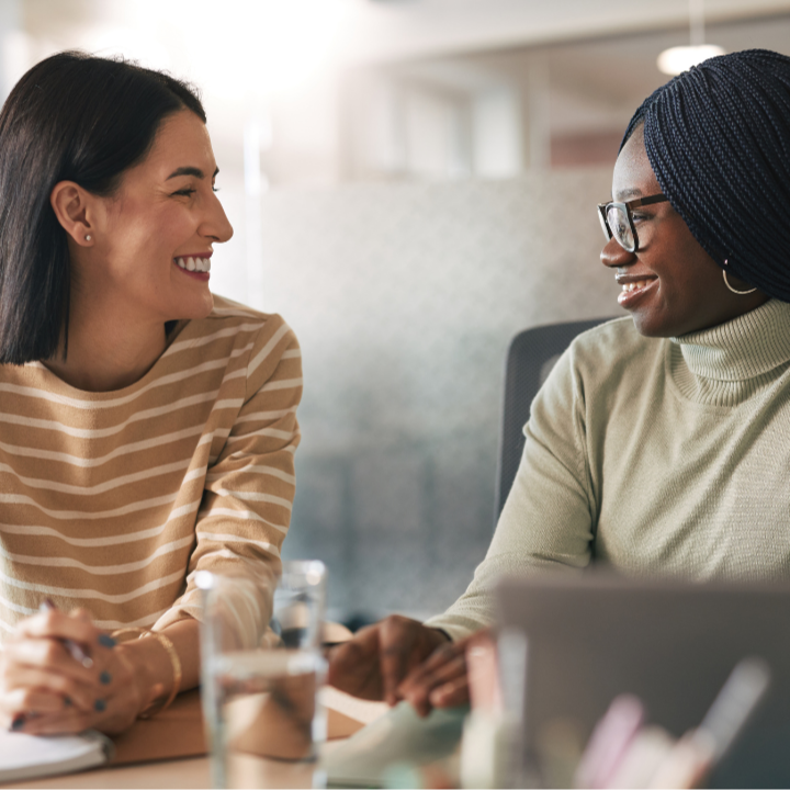 Two women sitting at a table in an office setting, smiling and engaged in conversation.