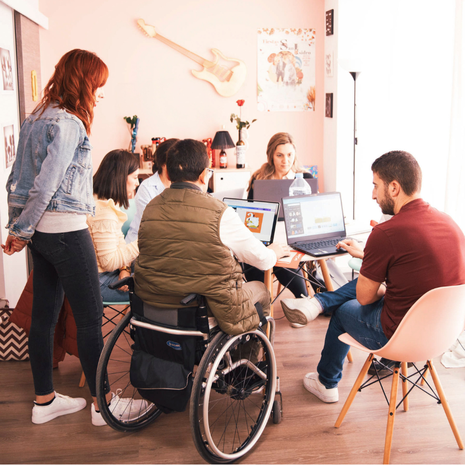 Group of people, including a person in a wheelchair, using laptops in a casual office setting.