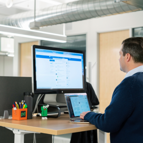 Man using a laptop at a desk with multiple computer monitors in an office setting.